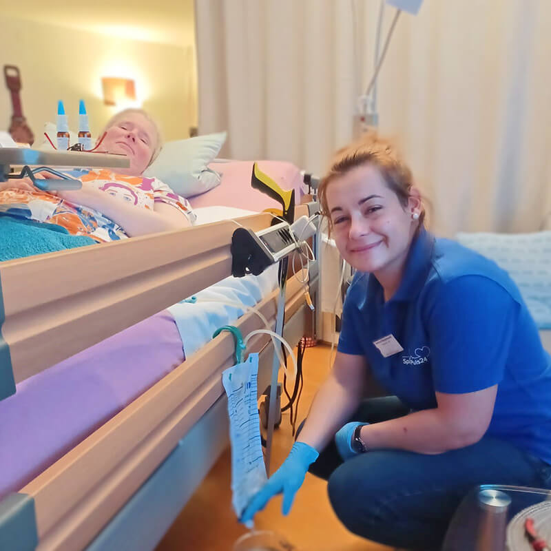 Pflegerin hockt freundlich lächelnd am Bett einer pflegebedürftigen Frau