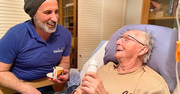 Pfleger füttert und unterhält sich mit im Bett liegendem Senioren