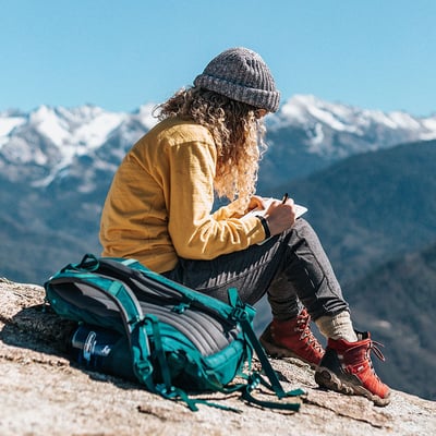 Frau im Wander-Outfit schreibt auf einem Berg etwas in ihr Heft