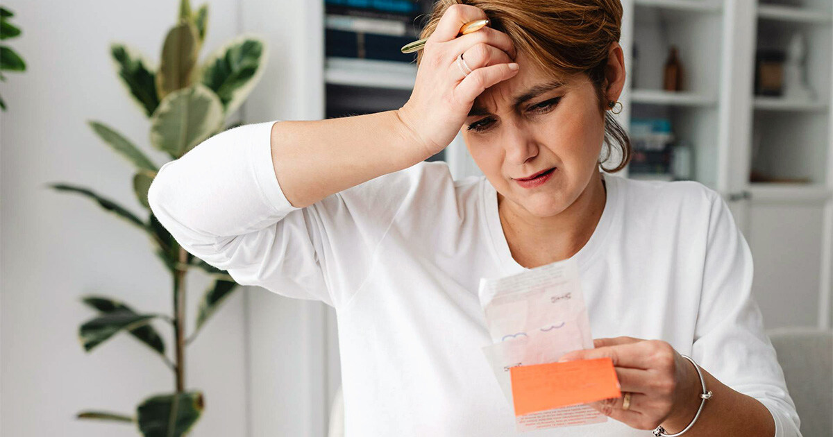 Besorgte Frau mit Kopfschmerzen liest eine Rechnung, während sie über die Kosten der Spitex nachdenkt, in einem modernen Wohnzimmer.