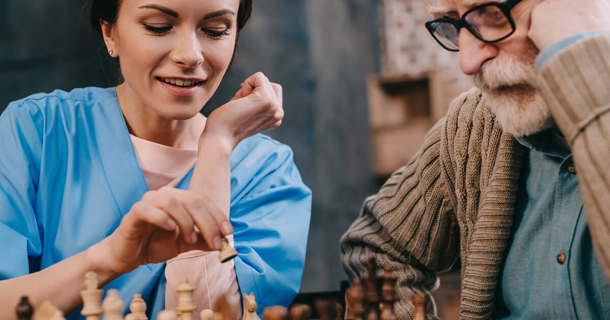 Jüngere Pflegerin spielt Schach mit älterem Mann im Rahmen der Seniorenbetreuung