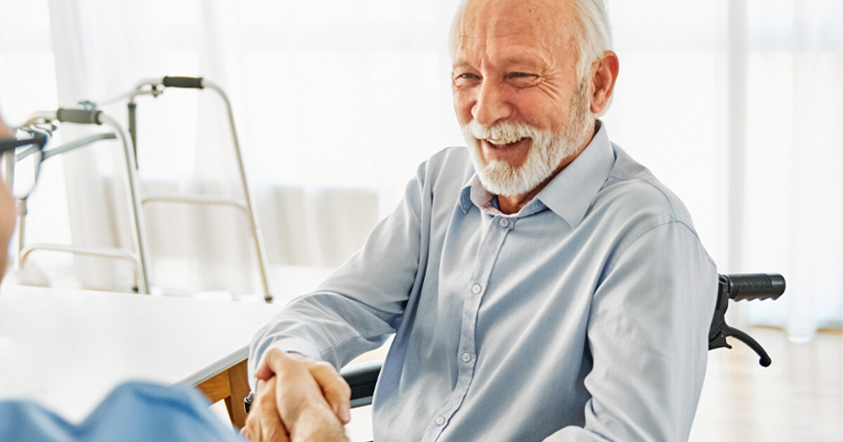 Lächelnder älterer Herr im Rollstuhl schüttelt die Hand eines Pflegedienstleiters in einer hellen Wohnung, im Hintergrund eine Gehhilfe.