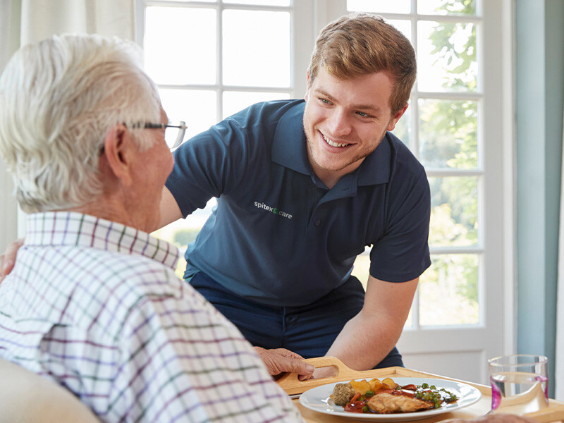 Ein Fachmann für Betreuung in einem blauen Shirt lächelt einen älteren Herrn an, während er ihm eine Mahlzeit serviert.