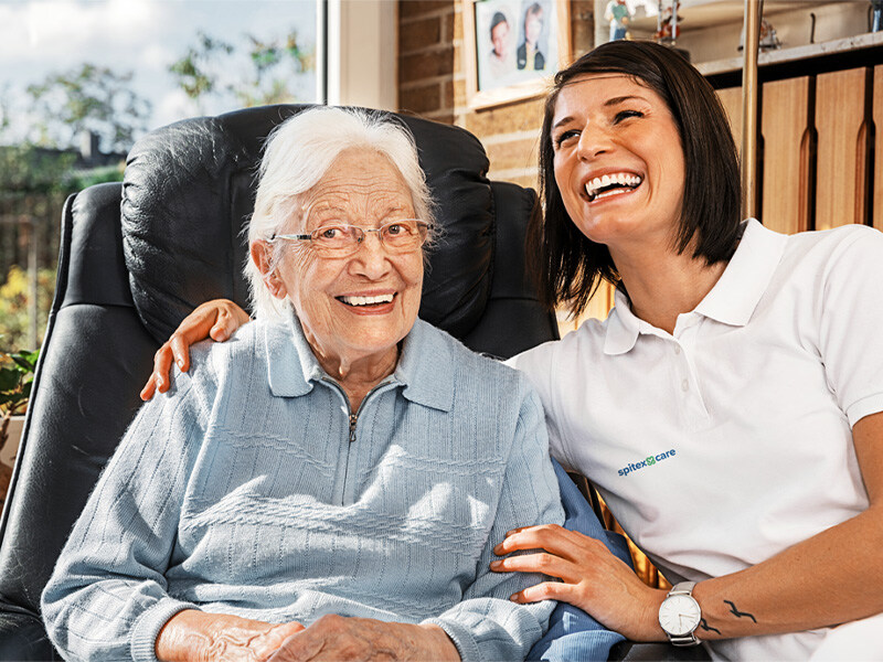 Fachfrau Gesundheit lacht zusammen mit einer älteren Dame, die im schwarzen Ledersessel sitzt, in einem hellen, wohnlichen Raum