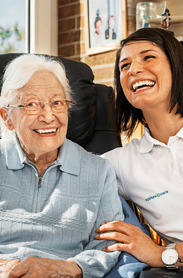 Fachfrau Gesundheit lacht zusammen mit einer älteren Dame, die im schwarzen Ledersessel sitzt, in einem hellen, wohnlichen Raum