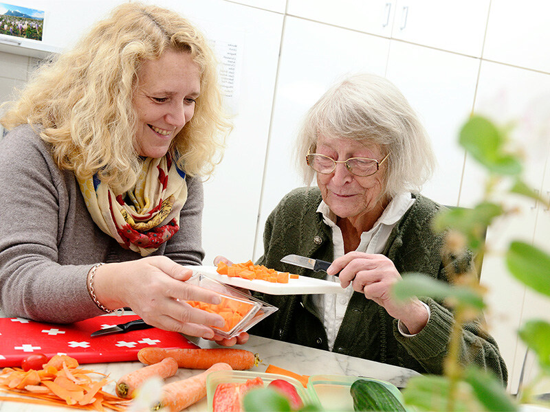 Eine hauswirtschaftliche Mitarbeiterin mit lockigen blonden Haaren hilft einer älteren Dame mit Brille beim Gemüseschneiden in einer Küche