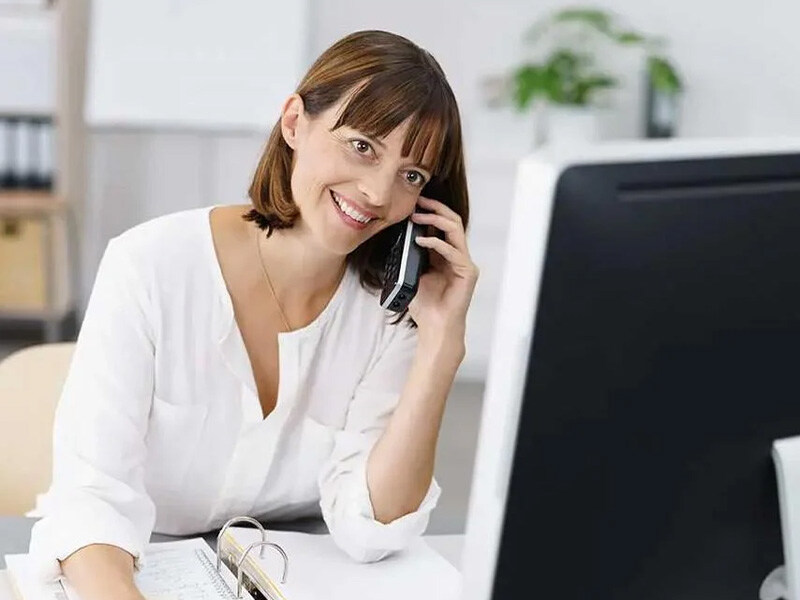 Freundliche Frau im Büro telefoniert, vor ihr ein Aktenordner und ein Bildschirm unscharf im Vordergrund