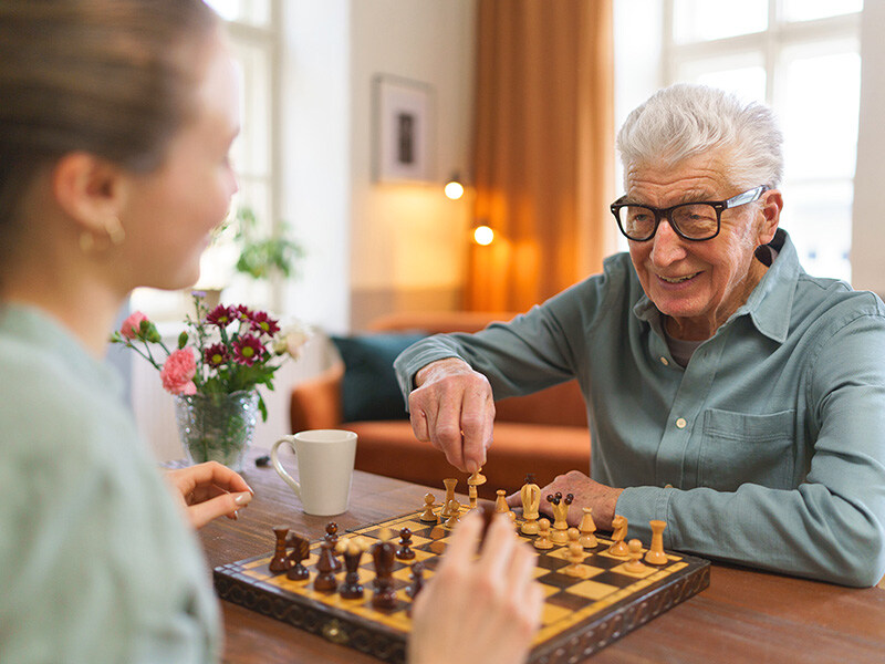 Herr mit Brille spielt Schach mit junger Frau im Wohnzimmer, eine Tasse Kaffee auf dem Tisch, Gemütlichkeit und Konzentration.