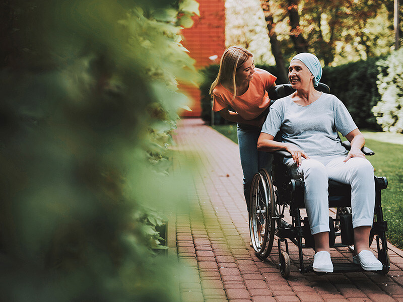 unge Frau schiebt lächelnde ältere Dame im Kopftuch im Rollstuhl durch einen grünen Park, ein Bild der Unterstützung in der Onkologiepflege.