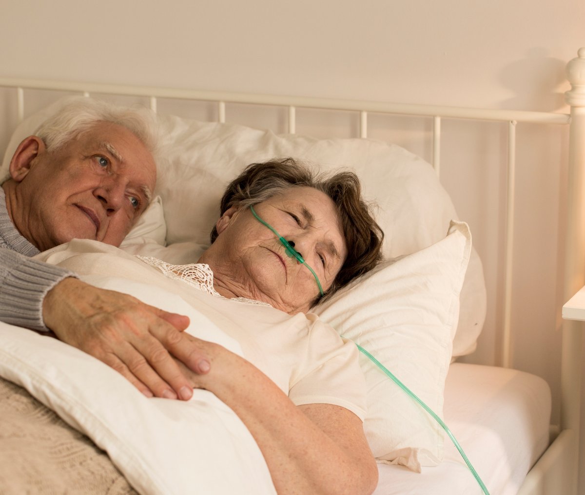 Älterer Mann liegt neben seiner schwer kranken Frau im Bett und sieht besorgt aus