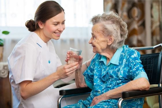 ältere Frau im Rollstuhl nimmt ein Glas Wasser aus de Hand ihrer Pflegerin