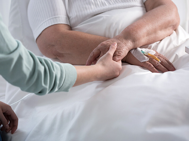 Eine Person hält tröstend die Hand einer älteren Person mit einem IV-Zugang im Bett, symbolisch für private Spitex-Dienste im häuslichen Umfeld.