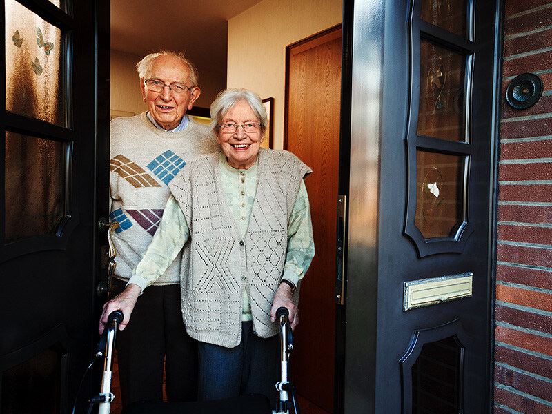 Ein älteres Paar steht lächelnd in der Türöffnung ihres Hauses, beide mit Gehhilfen. Sie stehen vor einer dunklen Haustür.