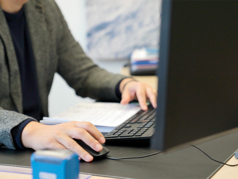 Pflegedienstleiter an einem Schreibtisch, bedient eine Tastatur und eine Computermaus, im Hintergrund sind Büro und Monitor unscharf zu erkennen.