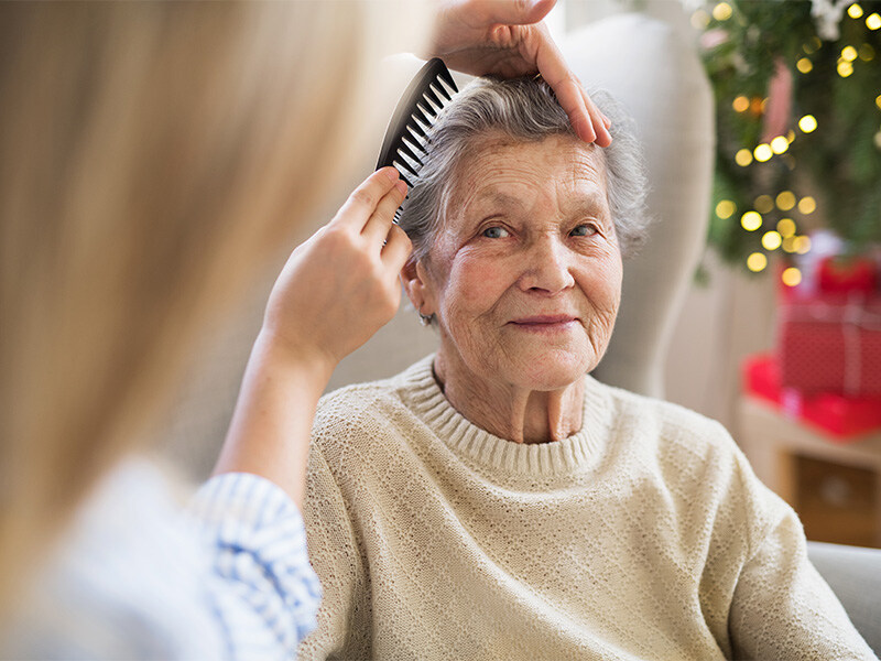 Eine Pflegehelferin kämmt liebevoll das Haar einer lächelnden älteren Frau, die in einem gemütlichen Raum mit Weihnachtsdekoration sitzt.