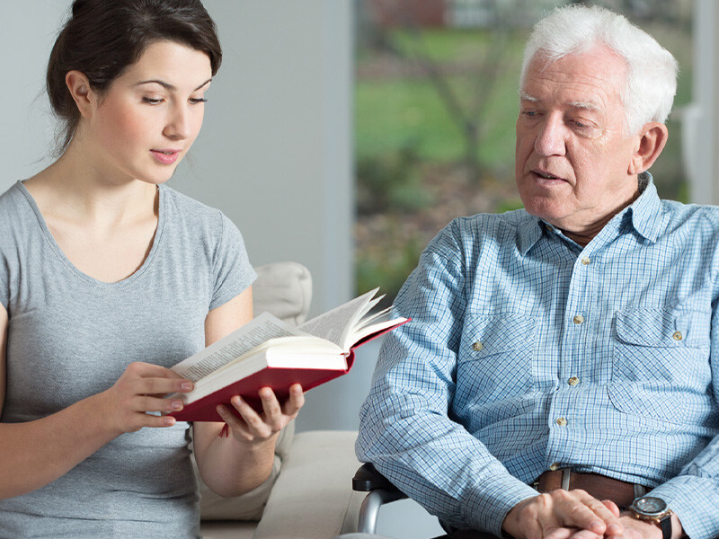 Eine junge Frau liest einem älteren Mann, der auf einem Rollstuhl sitzt, aus einem Buch vor, beispielhaft für das Pflegen von Eltern zu Hause.