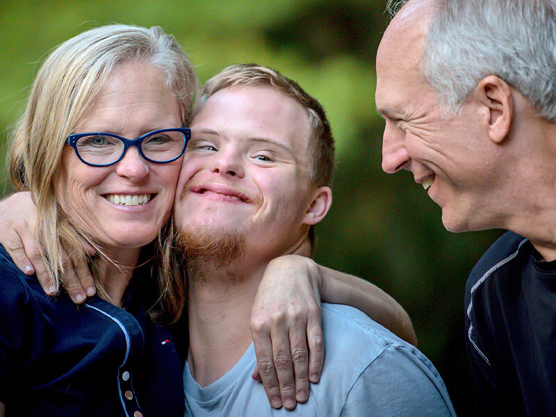 Lächelnde pflegende Angehörige umarmen liebevoll ihren erwachsenen Sohn mit Down-Syndrom im Freien.