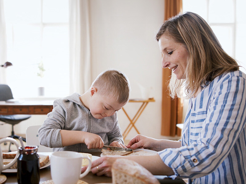 Mutter und Kind mit Downsyndrom frühstücken gemeinsam, Mutter hilft Kind beim Brotschmieren