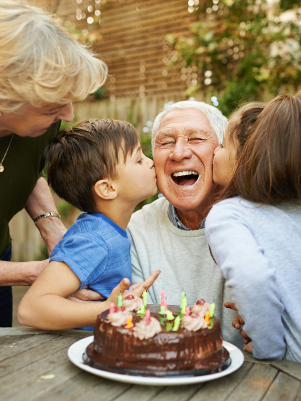 Glücklicher älterer Mann wird von seinen Enkelkindern auf einer Geburtstagsfeier im Freien geküsst, während eine ältere Frau zuschaut