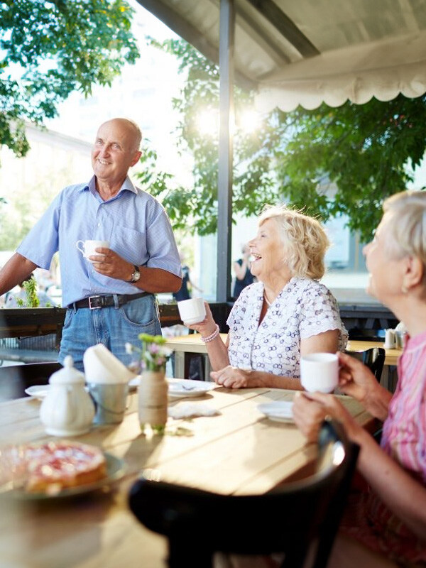 Drei Senioren lachen und unterhalten sich bei einem Kaffeekränzchen im Freien an einem sonnigen Tag