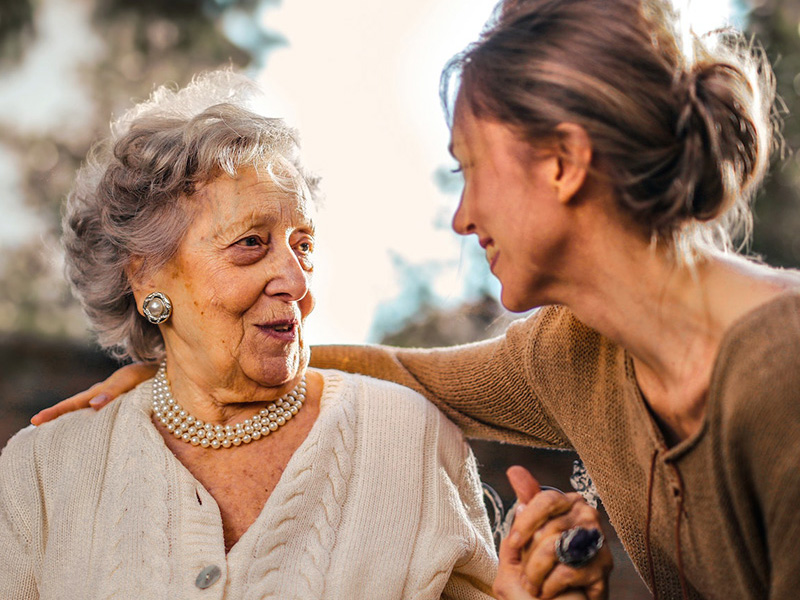 Eine ältere Dame mit Perlenkette lächelt dankbar, während sie von einer jüngeren Frau, vermutlich einer pflegenden Angehörigen, liebevoll umarmt wird.