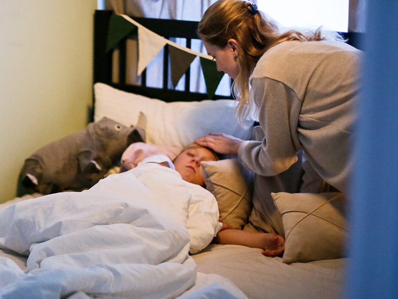 Eine junge Frau beugt sich fürsorglich über ein schlafendes Kind im Bett, ein Bild häuslicher Pflege durch Angehörige.