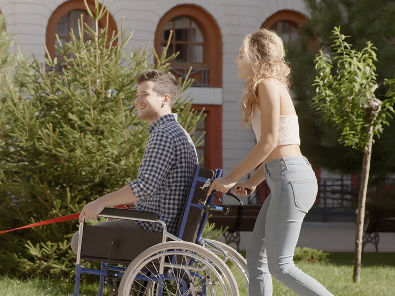 Junge Frau schiebt jungen Mann im Rollstuhl, Mann hält rote Leine, beide könnten ein Paar sein