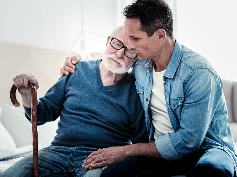 Ein älterer Herr mit Brille und blauem Pullover lehnt sich an Sohn, der ihn umarmt, in einem Moment der Fürsorge durch pflegende Angehörige.